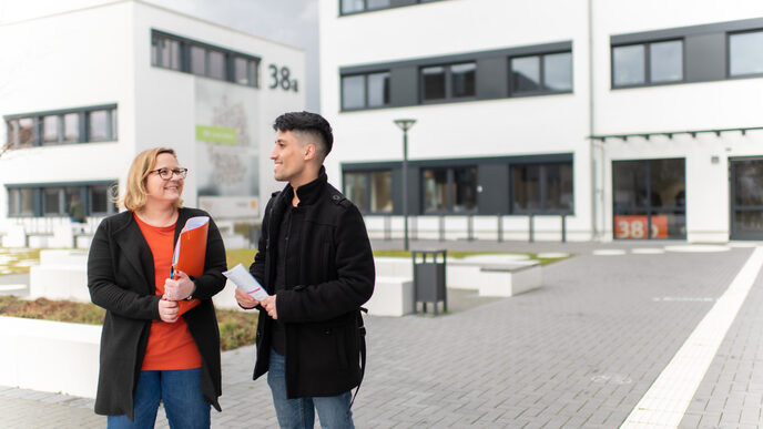 Photo of an employee standing with a student in front of a campus building. She is holding an orange folder under her arm, he has a flyer in his hand.
