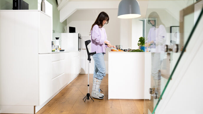 Eine weiblich gelesene Person steht in einer Küche. Sie hat den rechten Fuß ein einer Schiene. Neben ihr steht die Gehhilfe mit dem STEETS-Modul.