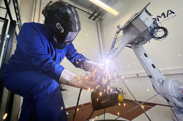 On the left, a man in a blue work suit with a welding helmet. He is welding the ends of several rods together. On the right, a robot arm holding one of the rods in position. In the middle is the welding spot with a shower of sparks.