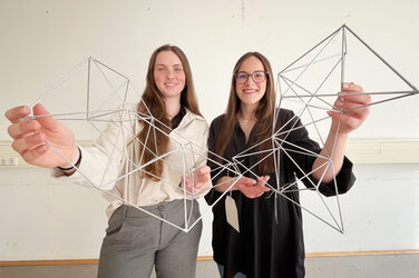 Two people smilingly hold a delicate structure made of metal rods up to the camera.