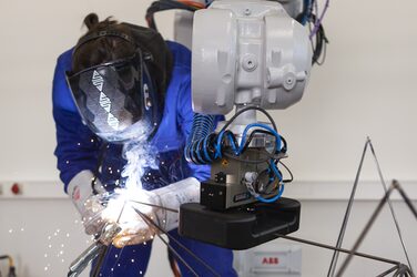 In the center foreground, part of a grey robot arm protrudes into the picture from above. The black clamp at the end of the arm holds a metal rod in position. Behind it, a person in a blue work suit and welding helmet can be seen bending over the other end of the rod and welding it to other rods.
