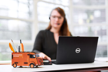 Foto einer Stiftebox in Form des Kastenwagens der Fachhochschule auf einem Schreibtisch. Unscharf dahinter sitzt eine Frau mit Headset am Laptop.