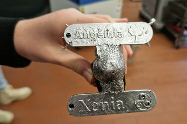 One hand holds the silver metal casting of two connected signs with the names Angelina and Xenia.