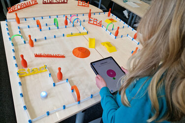 One person is holding a tablet. There is a ball on the table in front of them and an obstacle course is made up of several objects.