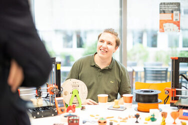 Ein Mitarbeiter des FabLabs der Fachhochschule Dortmund erklärt einer interessierten Person, wie der auf dem Tisch stehende 3D Druck funktioniert.An employee of the FabLab at Dortmund University of Applied Sciences and Arts explains to an interested person how the 3D printer on the table works.