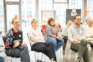 Acht Menschen im Bild hören den Info-Talks mit Kurzpräsentationen zu Auslandsaufenthalten zu und lachen dabei. Sie befinden sich im hellen großen Glasanbau des Mensafoyers am Standort Sonnenstraße. Eight people in the picture listen to the info talks with short presentations on stays abroad and laugh at the same time. They are in the bright, large glass extension to the cafeteria foyeron campus Sonnenstraße