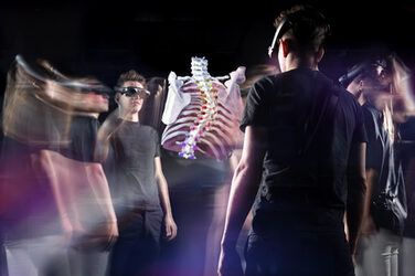 A group of young people standing in a circle look at the hologram of a human torso skeleton through virtual reality glasses.