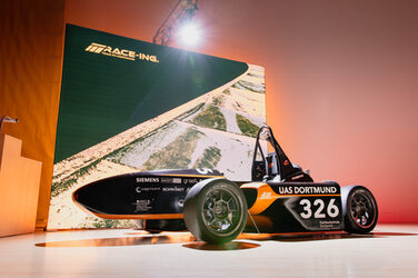 A racing car is parked on a stage, behind it a screen with the inscription "Race-Ing. Team Fachhochschule Dortmund".