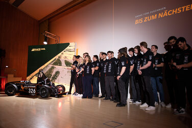 Around 20 people in black T-shirts are standing on a stage, with a racing car to their left.
