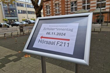 In einem Innenhof steht ein Hinweisschild für den Weg zum Hörsaal F211 am Schüler*innen-Tag.