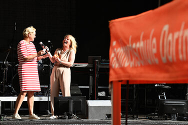 Zwei Personen auf einer Bühne, die linke spricht, die rechte lacht. Rechts vorn ragt die Ecke eines orangefarbenen Sonnendachs ins Bild.