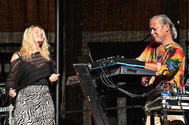 Two musicians on a stage, the person on the left has their eyes closed and appears to be dancing, the person on the right operates several keyboards and other devices.