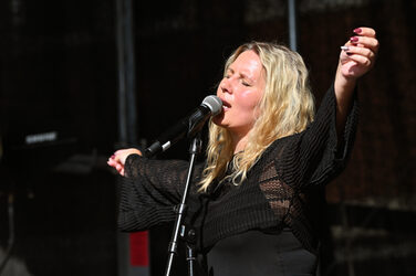 Eine Person mit langen blonden Haaren und ausgestreckten Armen singt mit geschlossenen Augen in ein Mikrofon.