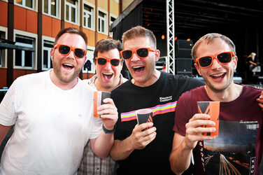 Four people with orange sunglasses and drinking cups in their hands laugh into the camera.