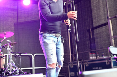 A man in fashionably torn jeans on a stage grins exaggeratedly into the camera.