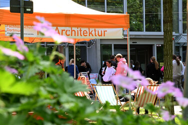 Durch violette Blüten im Vordergrund ist ein orangefarbenes Zelt mit der Aufschrift "Architektur" zu sehen, daneben mehrere Menschen vor einem Gebäude.