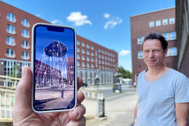 In the foreground on the left, a hand holds a cell phone in the picture, showing the same place as in the background, but with a virtual sculpture on it. In the foreground on the right is a person in a gray T-shirt.