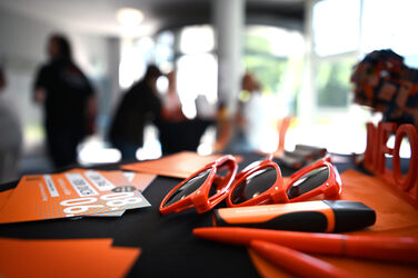 Orange sunglasses, pens and postcards lie on a black surface, several people can be seen blurred in the background.