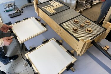 Studierende platzieren Objekte in einem Muster auf zwei quadratischen Lochblechen auf einem Tiefziehtisch aus Holz. Gleichzeitig werden weiße Polystyrolplatten mithilfe von Klammern in einem quadratischen Holzrahmen stabilisiert.