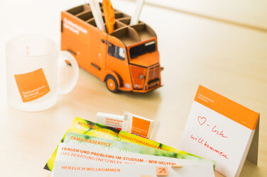 Photo of a still life with various flyers, an FH mug, the HY pencil box and a notepad with "Welcome" written on it.