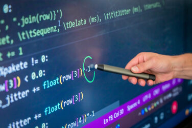 Photo of a section of a digital board on which a code can be seen. Someone is holding a pen pointing to a spot.