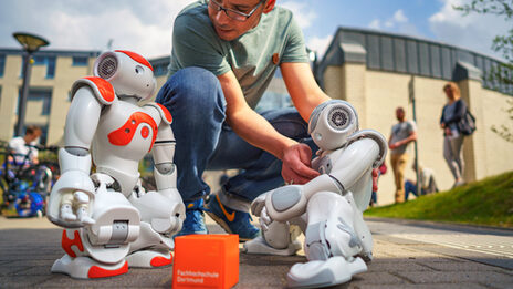 Photo of two NAO robots standing outside Building 42. An employee sets up the robots. A UAS cube lies between the robots.