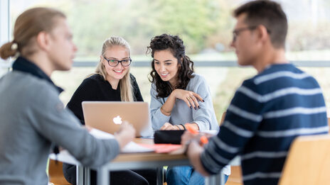 Foto von vier Studierenden an einem Gruppentisch. Im Fokus zwei Studentinnen, die nebeneinander sitzen und auf einen Laptop schauen. Zwei weitere Studenten sind in ein Gespräch vertieft.