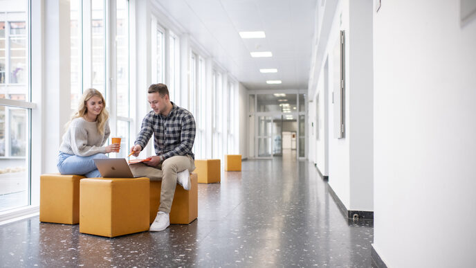 Foto von einer Studentin und einem Studenten. Sie sitzen im Flur in der FH und unterhalten sich. Der Student zeigt auf etwas am Laptop.