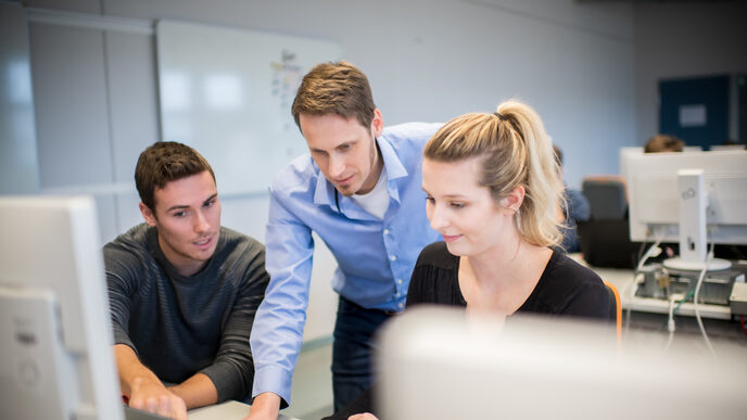 Foto eines Lehrenden im Computerraum, der zwischen einem Studenten und einer Studentin steht und beiden etwas am Laptop erklärt. __ <br>The professor stands between a male student and a female student in the computer room and explains something to both of them on the laptop.