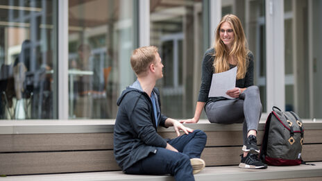 Photo of a male and a female student sitting on wooden steps and talking on campus. The female student is holding a note.__One male and one female student sitting on wooden steps and talking, the female student is holding a note.