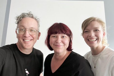 Drei Personen vor einer grauen Wand lächeln breit in die Kamera.