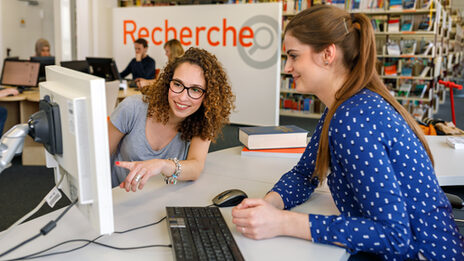 Photo of a student showing a library employee something with her finger on the monitor. In the background, a part of the library and people at computers can be seen out of focus.