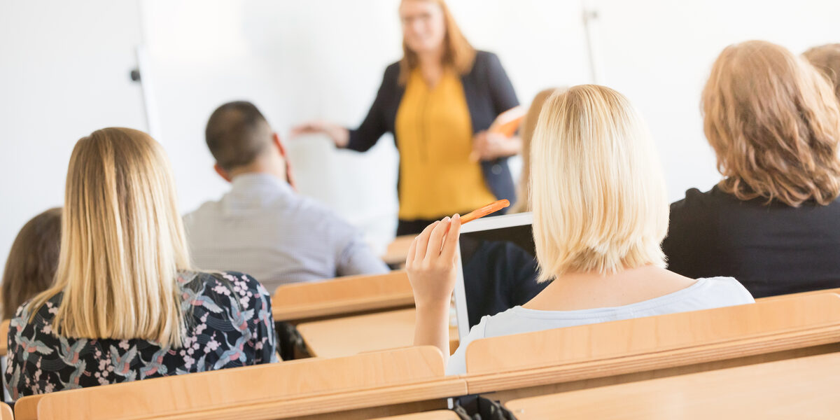 Foto von Hinterköpfen mehrerer Studierender in Sitzreihen im Hörsaal. Vorne ist eine Dozentin unscharf zu erkennen.