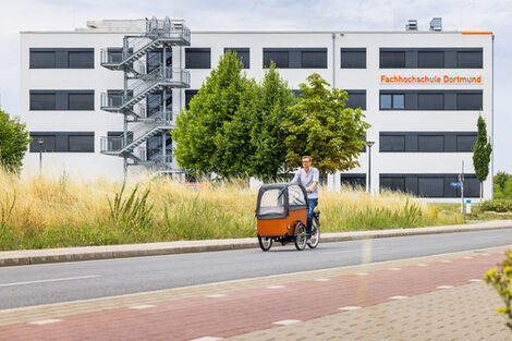 Photo of a man on a cargo bike riding away from a building on the street. The words "Fachhochschule Dortmund" can be seen on the building.
