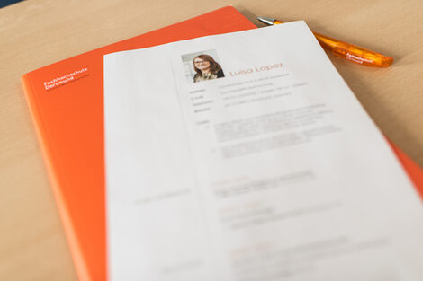 Photo of an orange-colored folder and application documents or a résumé on it on a table. __ Orange-colored folder and application documents or a résumé on it on a table.