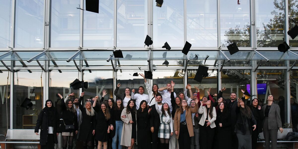 The graduates stand in front of the faculty building and throw their graduation hats in the air.