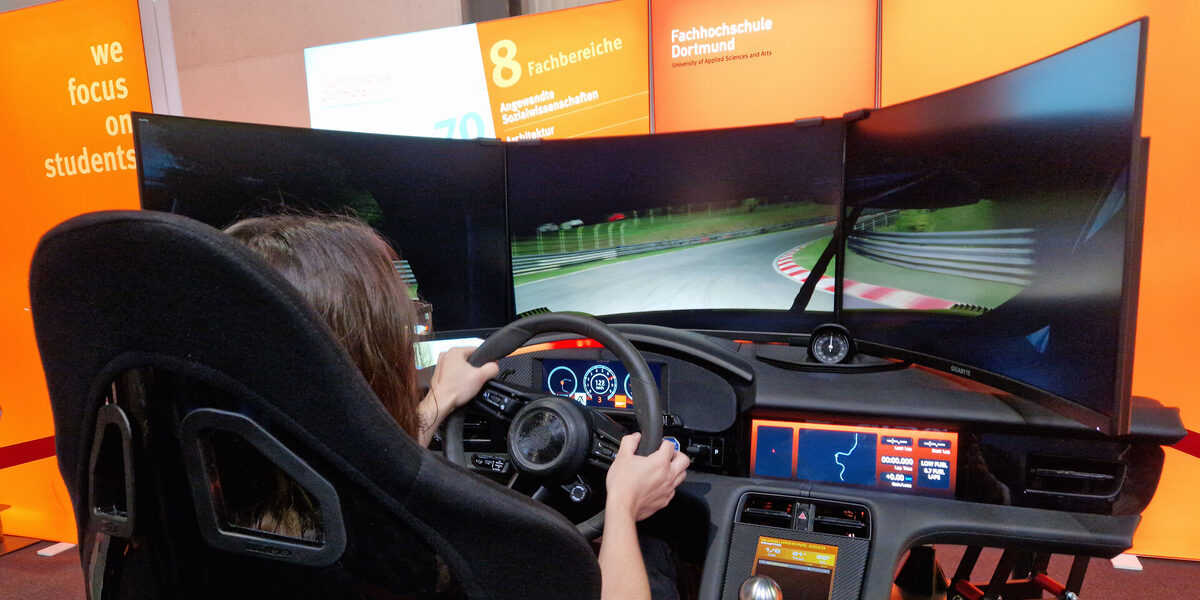 One person sits in the driving simulator, controls the steering wheel and looks at three screens.