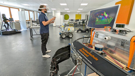 A person wearing VR goggles stands in a laboratory. In front of her is a monitor with colorful lines.