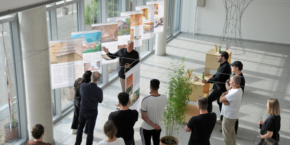 Blick in die im Foyer des Fachbereichs aufgebaute Ausstellung "Fibra Award Bambus", mit einer Gruppe Studierender, die der Vorstellung von Andrès Bäppler zuhören.