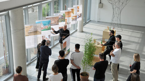 Blick in die im Foyer des Fachbereichs aufgebaute Ausstellung "Fibra Award Bambus", mit einer Gruppe Studierender, die der Vorstellung von Andrès Bäppler zuhören.