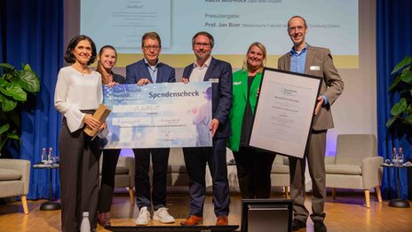 Six people stand next to each other on a stage. They present a large-format donation check and a certificate.