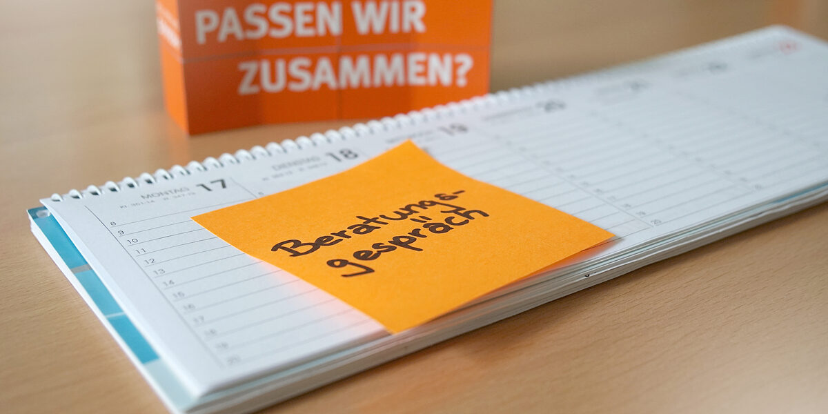 Foto eines Tischkalenders, auf dem ein orangefarbenes Post-It mit der Notiz „Beratungsgespräch“ klebt. Im Hintergrund stehen gestapelte Würfel, auf denen „Passen wir zusammen?“ steht.