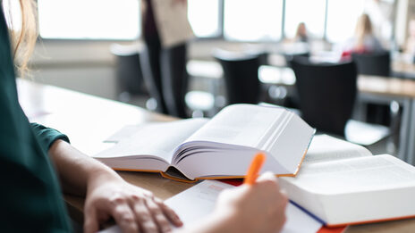 Foto von einer Frau, die sich Notizen macht. Vor ihr liegen zwei Bücher. Im Hintergrund befinden sich mehrere Leute im Seminarraum. __ Woman takes notes. There are two books in front of her. In the background there are several people in the seminar room.