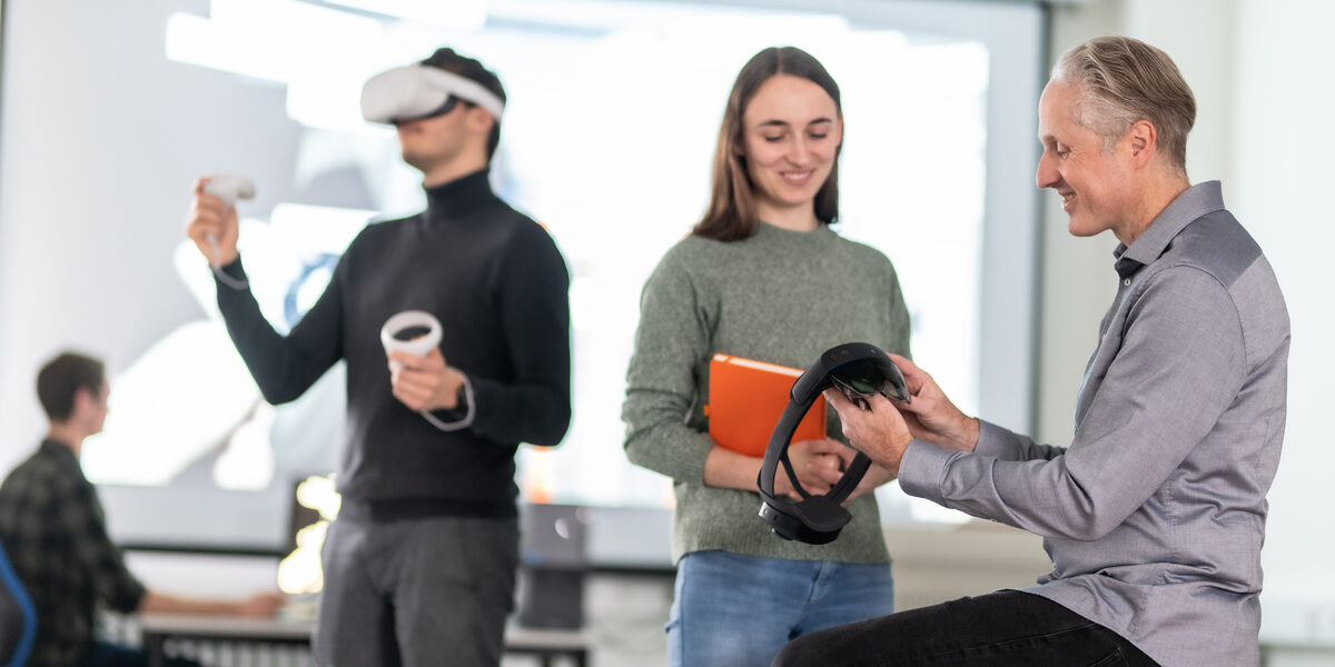Foto von einem Lehrenden und einer Studierenden. Der Lehrende zeigt der Studierenden eine VR-Brille. Zentral sieht man außerdem einen Studierenden, der eine VR-Brille trägt und diese bedient. Im Hintergrund sitzt ein Studierender an einem PC vor einer großen Leinwand.