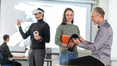 Foto von einem Lehrenden und einer Studierenden. Der Lehrende zeigt der Studierenden eine VR-Brille. Zentral sieht man außerdem einen Studierenden, der eine VR-Brille trägt und diese bedient. Im Hintergrund sitzt ein Studierender an einem PC vor einer großen Leinwand.