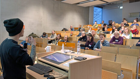 A male read person speaks to people in a lecture hall.