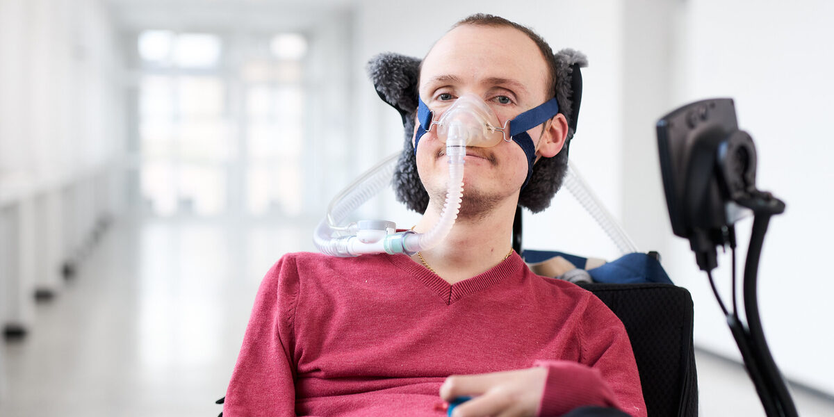 A person wearing a breathing mask has placed his hand on the control stick of the wheelchair in which he is sitting. A bright corridor can be seen in the background.
