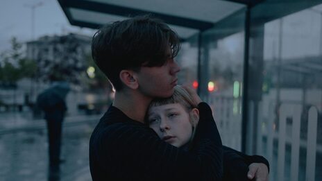 Two young adults embrace each other at a bus stop. The blurred background shows a street scene in rainy twilight.