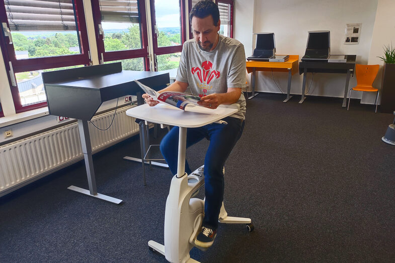 In the middle of a room is a bicycle-like device, albeit without wheels and handlebars, but with a small table. A person sits on the device, pedals and reads a magazine.