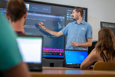 Photo of a lecture situation in a seminar room. A man is standing in front of a digital board and explaining something in code.